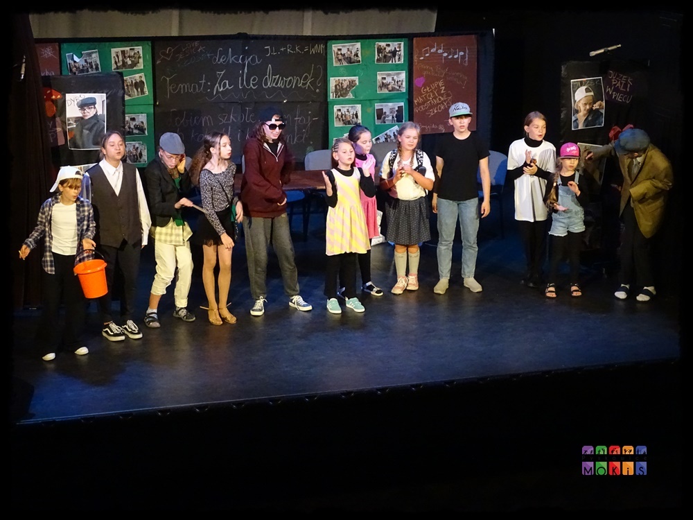 Zdjęcie przedstawiające grupę aktorów stojących w jednym rzędzie na scenie. W tle tablica lekcyjna z napisami oraz obrazki powieszone na zielonej ścianie