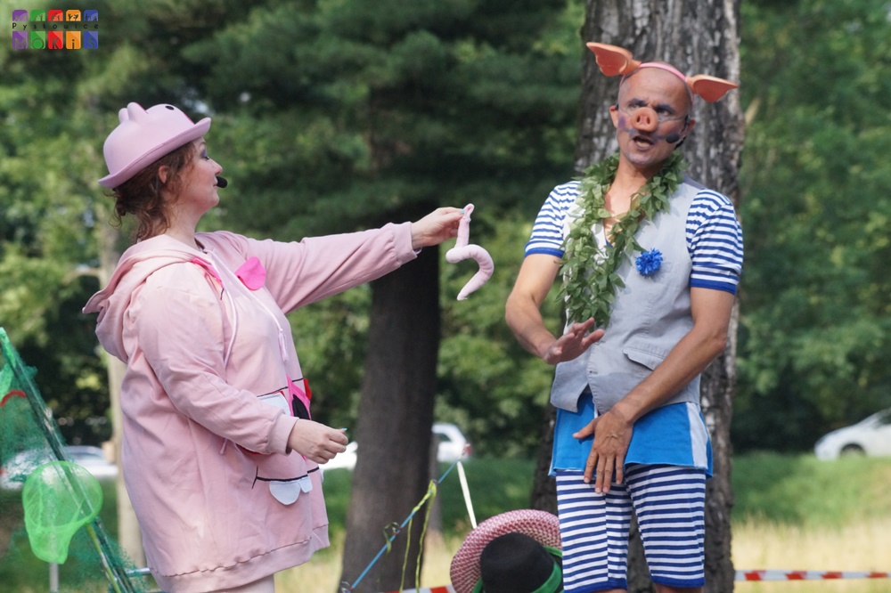 Zdjęcie przedstawiające kobietę przebraną za świnkę oraz mężczyznę przebranego za świnkę. Kobieta podaje świński ogon mężczyźnie. W tle widać drzewa parkowe.