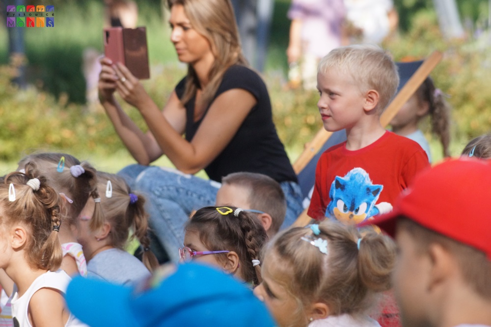 Zdjęcie przedstawiające patrzące dzieci w lewą stronę. W tle widać kobietę trzymającą telefon w rękach najprawdopodobniej robi zdjęcie.