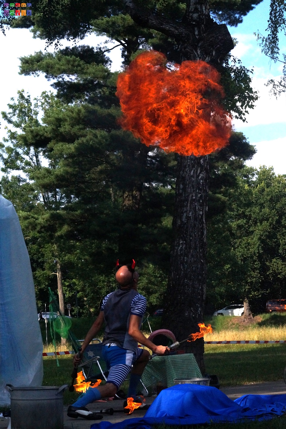 Zdjęcie przedstawiające mężczyznę patrzącego w górę przebranego w diabła. Nad nim unosi się ogień. W tle widać drzewa parkowe, trawnik