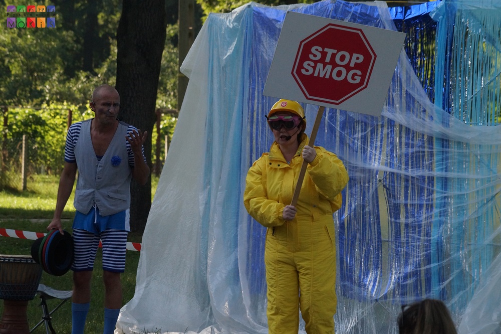 Zdjęcie przedstawiające kobietę w żółtym kombinezonie z tablicą w ręku z napisem stop smog. W tle po lewej stronie stoi mężczyzna ubrany w szorty w biało niebieskie paski. W tle widać scenografię z folii oraz drzewa parkowe.