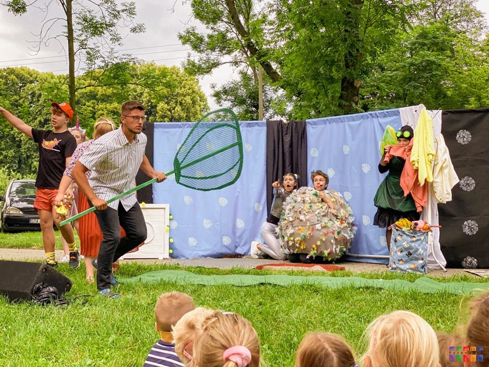 Zdjęcie przedstawia grających aktorów na scenie ustawionej na trawniku. W tle widać scenografię oraz drzewa parkowe.