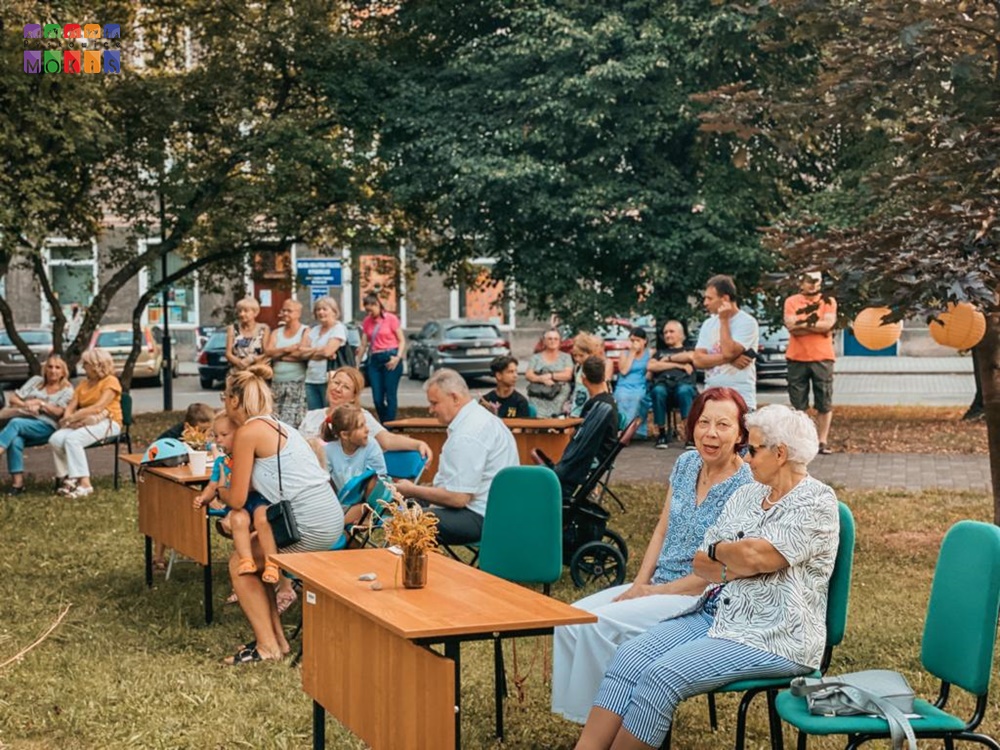 Zdjęcie przedstawia ludzi siedzących na krzesłach przy stołach w parku. W tle widać drzewa parkowe a w oddali blok mieszkalny.