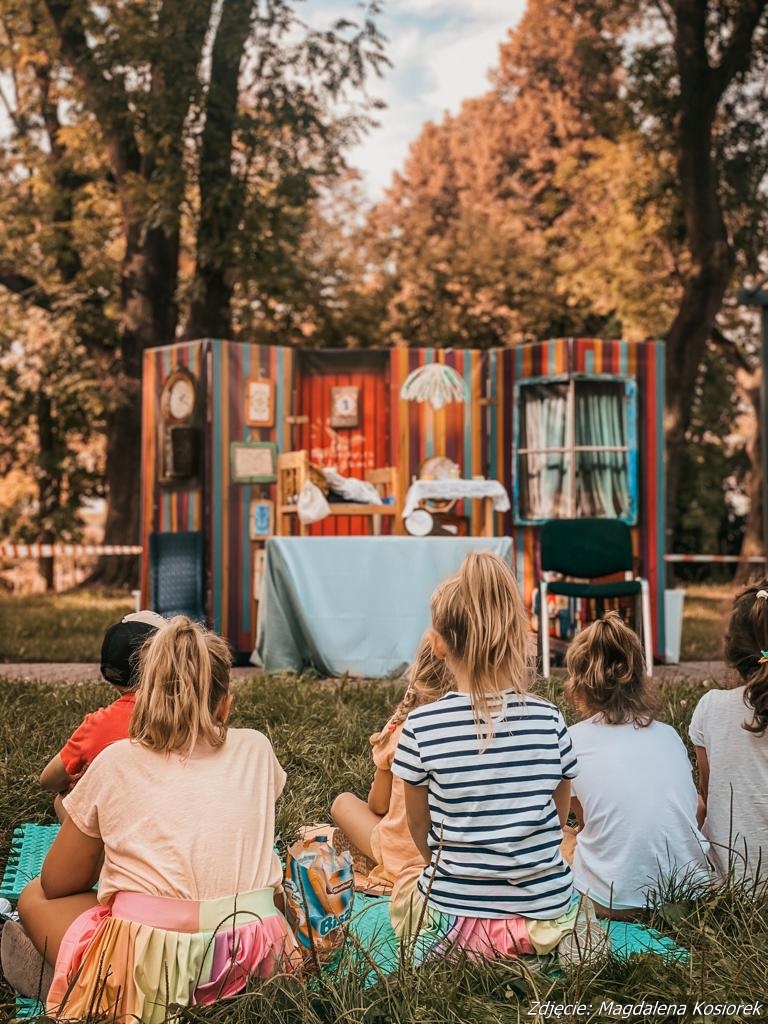 Zdjęcie przedstawiające siedzące dzieci na trawniku w parku i patrzące w kierunku sceny teatralnej.