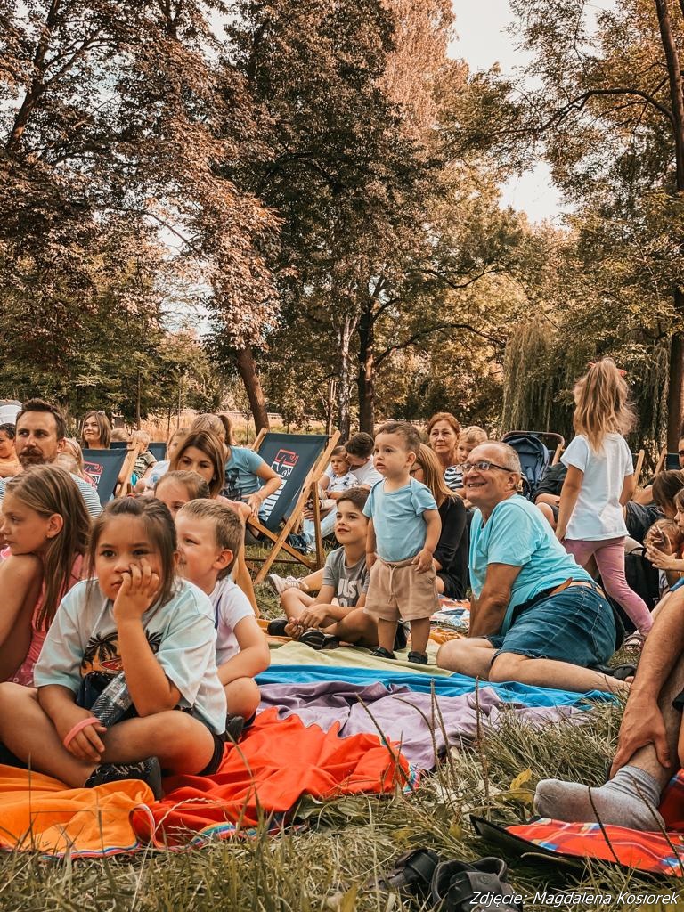 Zdjęcie przedstawia siedzące dzieci na kocach w parku. W tle widać drzewa parkowe.
