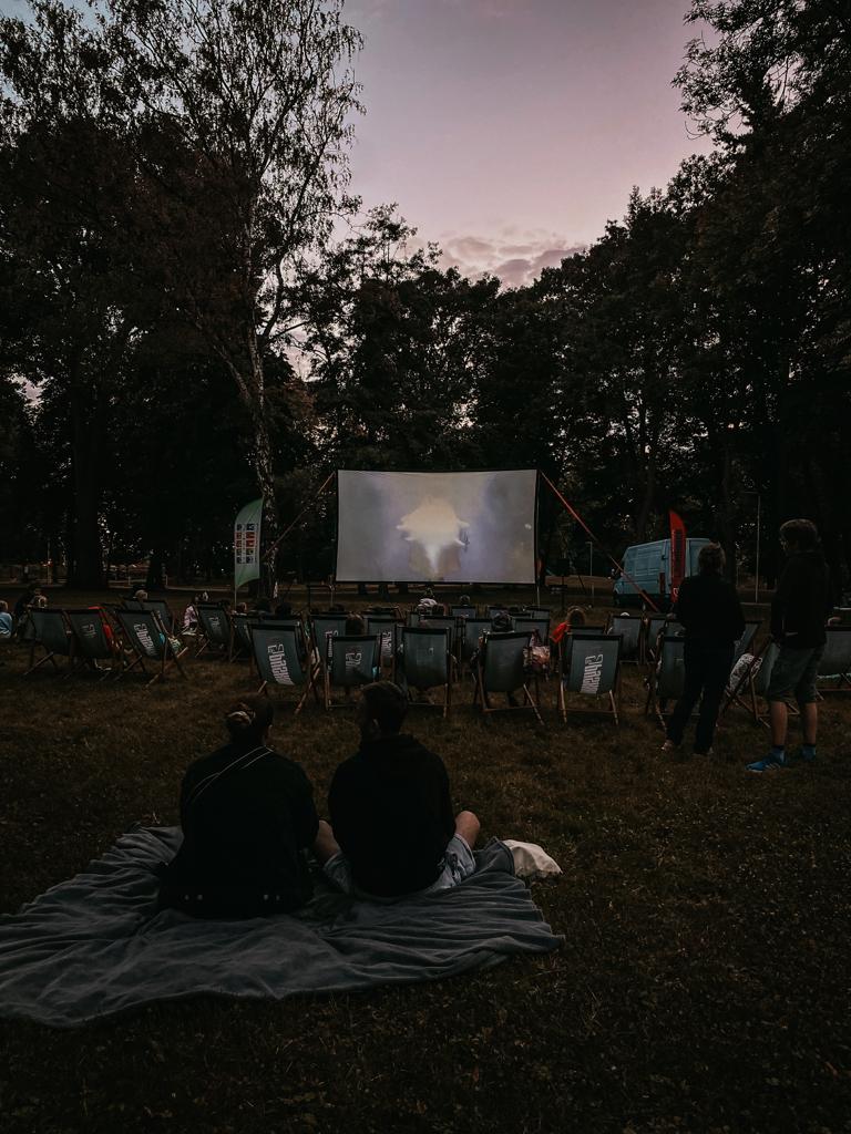 Zdjęcie przedstawia siedzących ludzi na leżakach obróconych w kierunku dużego ekranu, na którym wyświetlany jest film. W tle widać drzewa parkowe.