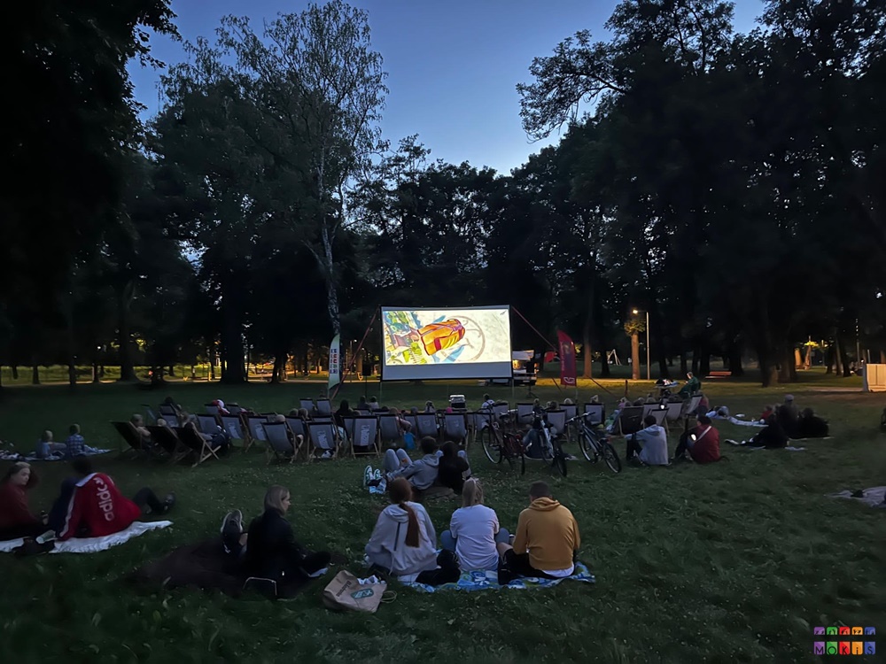 Zdjęcie przedstawiające siedzących ludzi na trawniku w parku i oglądającym na dużym ekranie film. W tle widać drzewa parkowe.