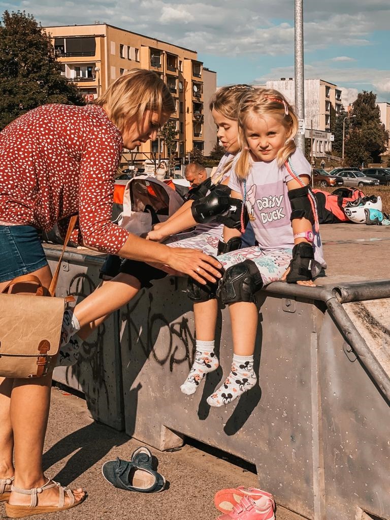Zdjęcie przedstawia siedzące dzieci na skateparku i ubierające z dorosłą osobą rolki