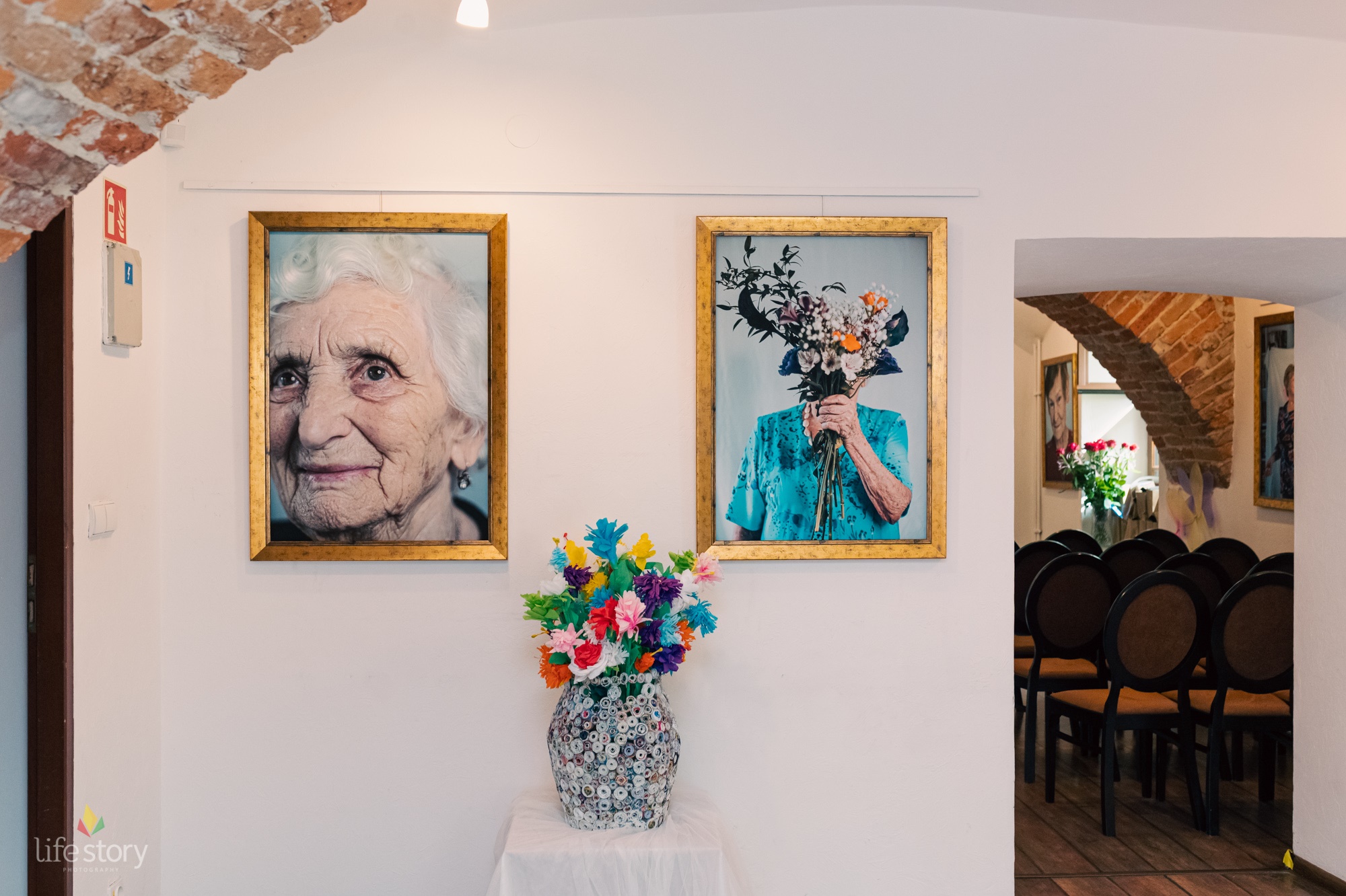 Zdjęcie przedstawia wiszące obrazy w galerii. W środkowej części na stoliku stoi dzban z kwiatami. Po prawej stronie przez drzwi widać ustawione krzesła.