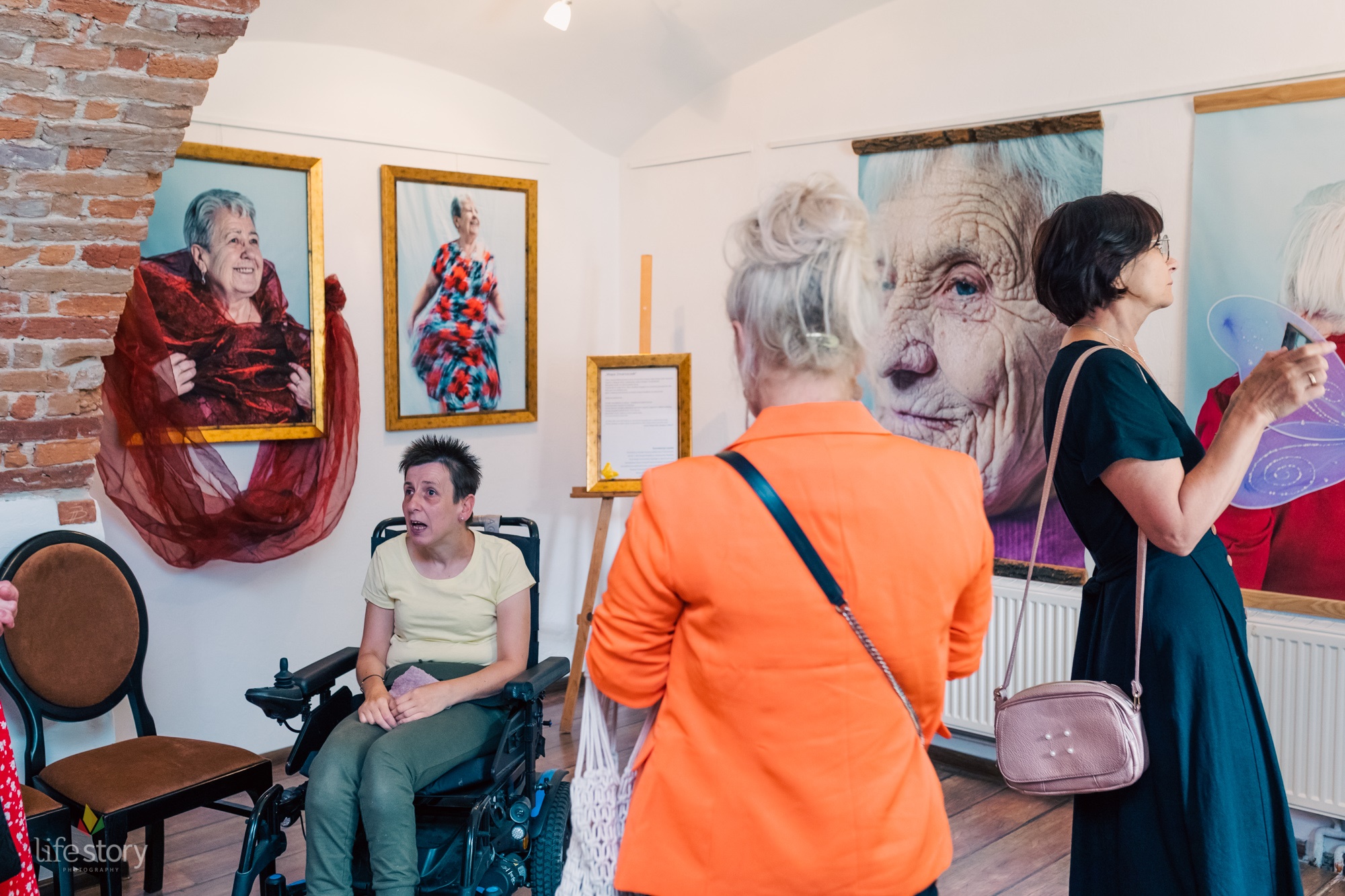 Zdjęcie przedstawia trzy kobiety oglądających wystawę obrazów, zdjęć w galerii. Jedna z nich siedzi na wózku inwalidzkim