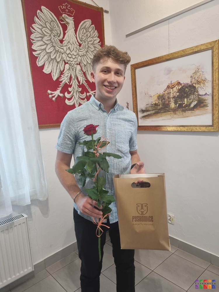 Zdjęcie przedstawiające mężczyznę stojącego i uśmiechającego się. W ręku trzyma różę oraz papierową torbę z napisem Pyskowice.