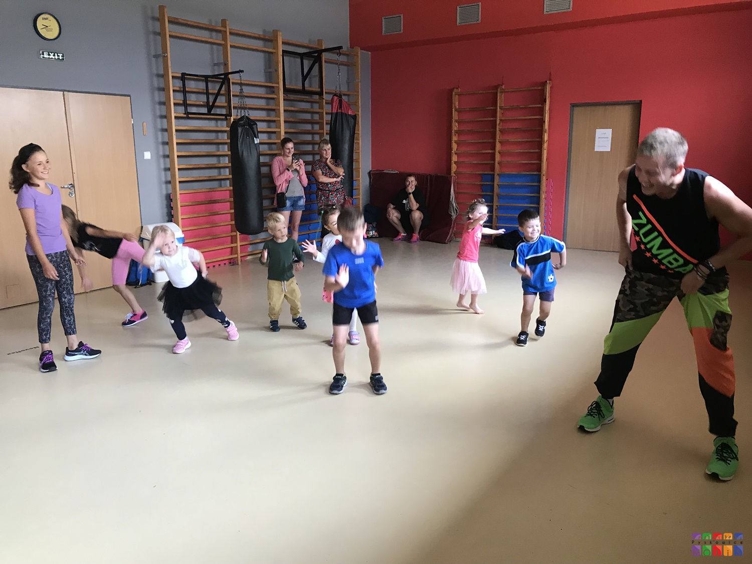 Zdjęcie przedstawiające tańczące dzieci w salce fitness. W tle widać drabinki oraz materace