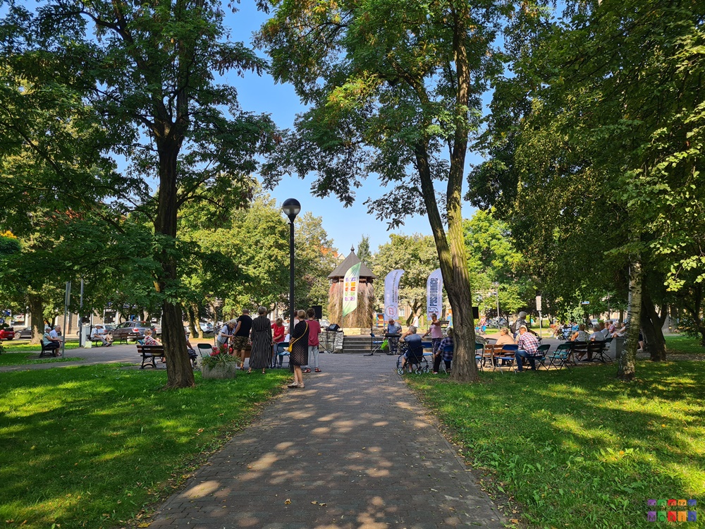 Zdjęcie przedstawia bawiących siedzących oraz stojących ludzi w parku. W środkowej jej części widać grającego Starszego Pana na organach