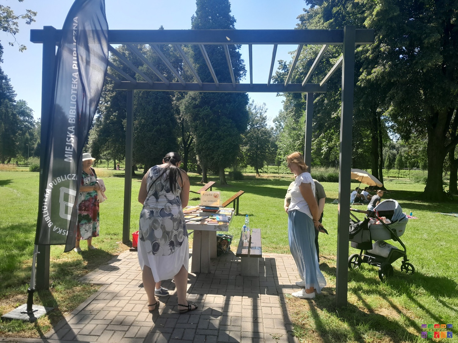 Zdjęcie przedstawiające stolik pod pergolą metalową przy którym stoją ludzie. W tle widać trawnik oraz drzewa parkowe. Po prawej wózek dziecięcy