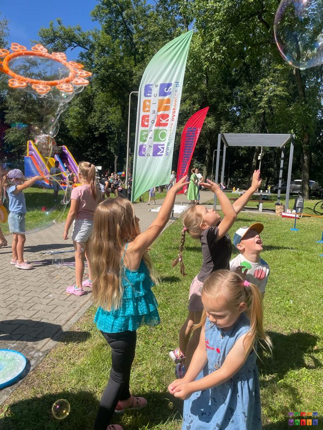 Zdjęcie przedstawiające bawiące się dzieci w parku, puszczają bańki mydlane. W tle widać powiewające flagi, dmuchany zamek kolorowy oraz pergola