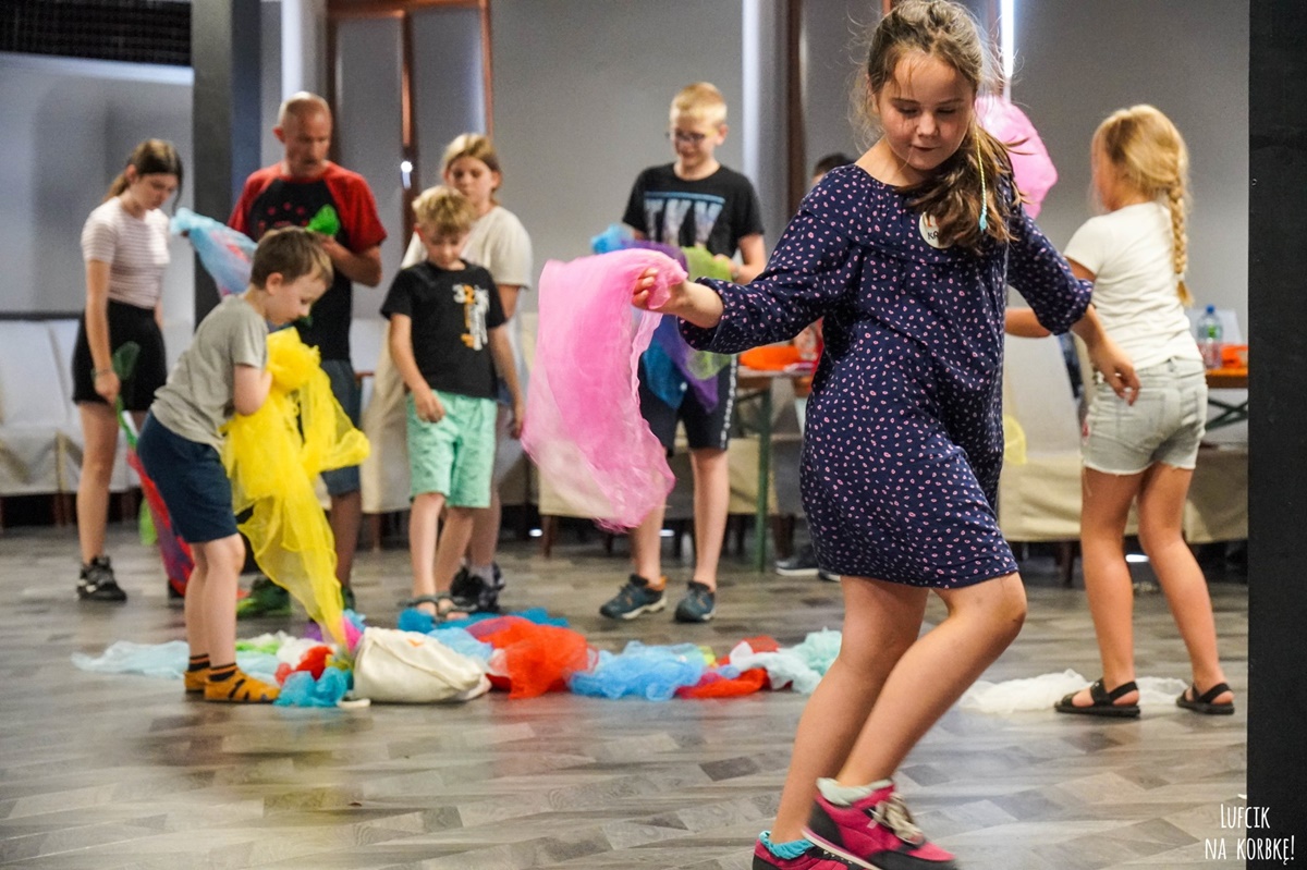 Zdjęcie przedstawia tańczącą z chustą dziewczynkę w warkowczu. W dalszej części zdjęcia widać dzieci z kolorowymi chustami