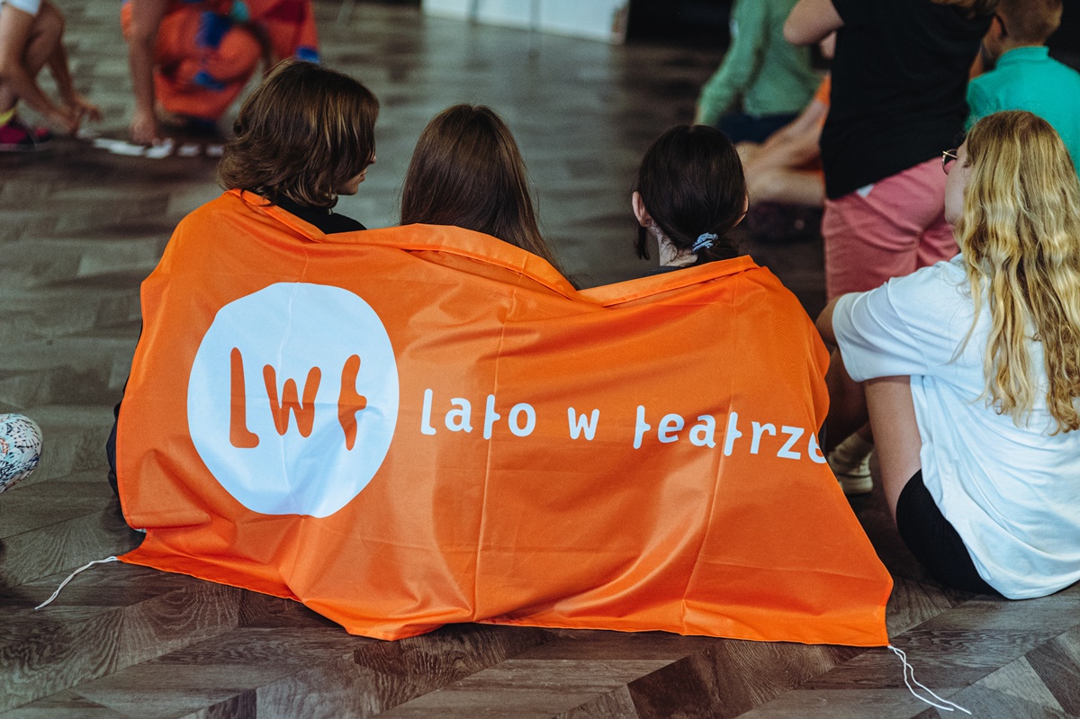 Zdjęcie przedstawiające siedzące trzy osoby a na ich plecach baner pomarańczowy z napisem lato w teatrze