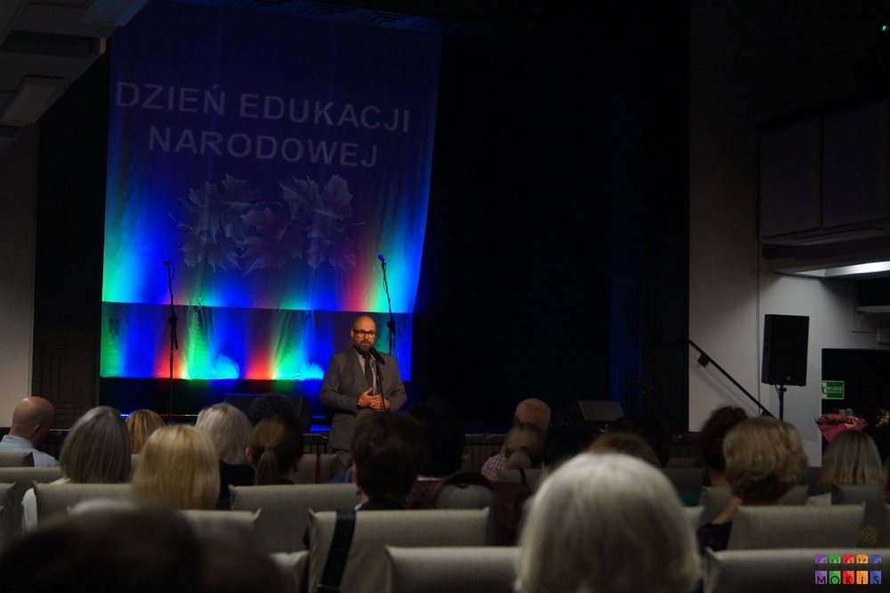 Zdjęcie przedstawia widownię siedzącą tyłem do fotografa. W oddali widać przemawiającego mężczyznę oraz scenę z napisem Dzień Edukacji Narodowej.