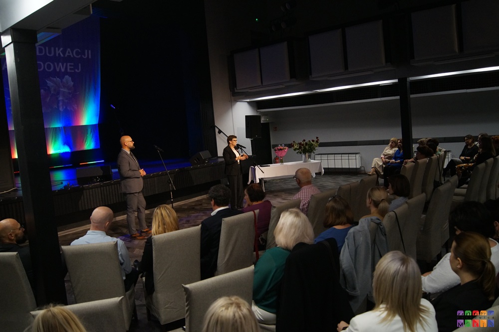 Zdjęcie przedstawiające z prawej strony widownię a sali widowiskowej z siedzącymi ludźmi a z lewej dwie osoby mówiące do mikrofonu na statywie