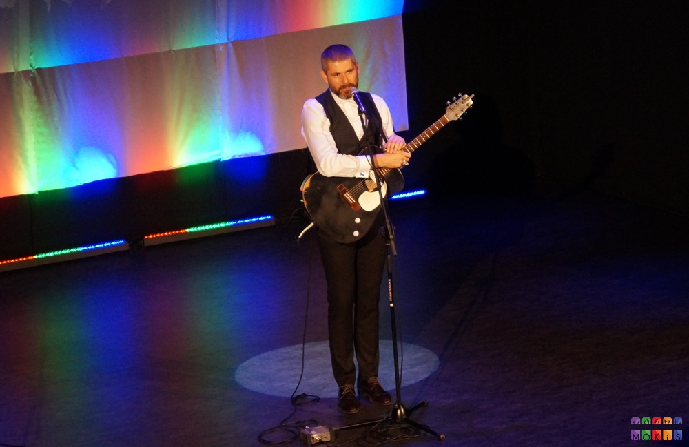 Zdjęcie przedstawia mężczyznę z gitarą stojącego przy mikrofonie na statywie. Tło kolorowe - tęczowe.