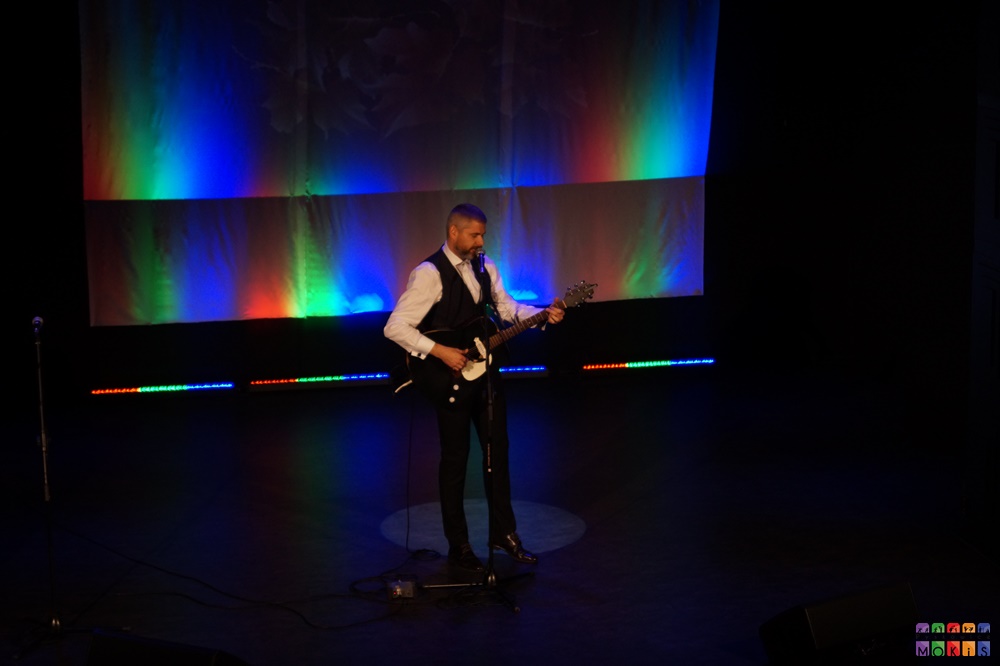 Zdjęcie przedstawia mężczyznę z gitarą stojącego przy mikrofonie na statywie. Tło kolorowe - tęczowe