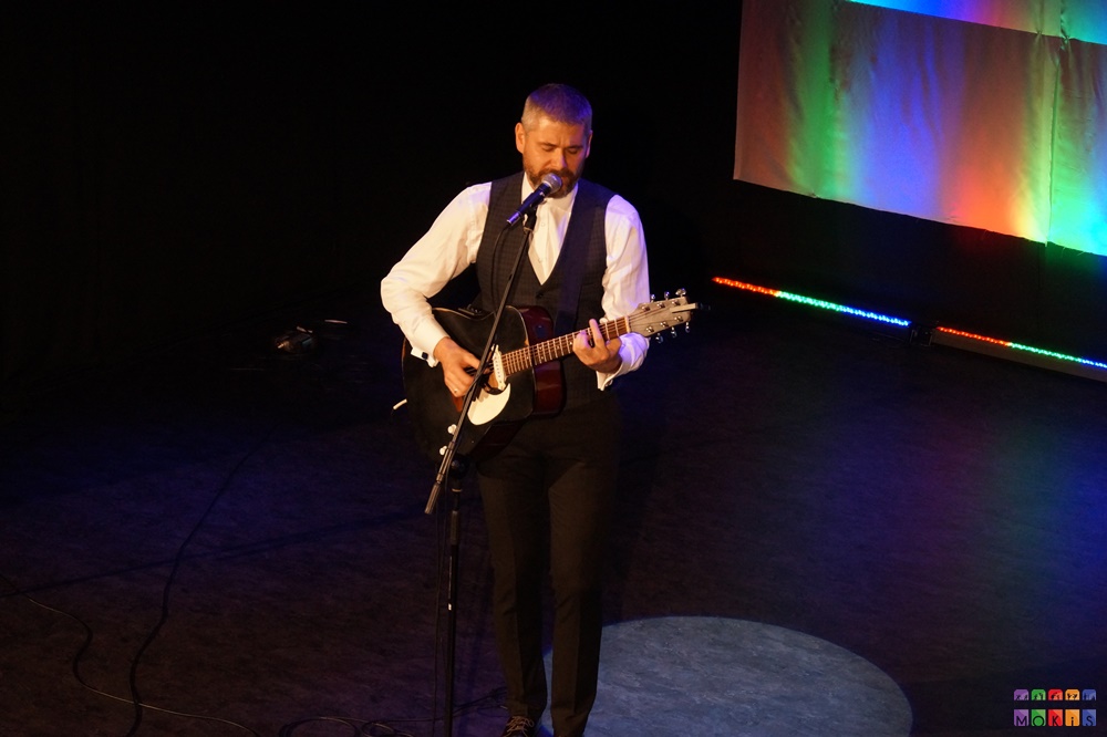 Zdjęcie przedstawia mężczyznę z gitarą stojącego przy mikrofonie na statywie. Tło kolorowe