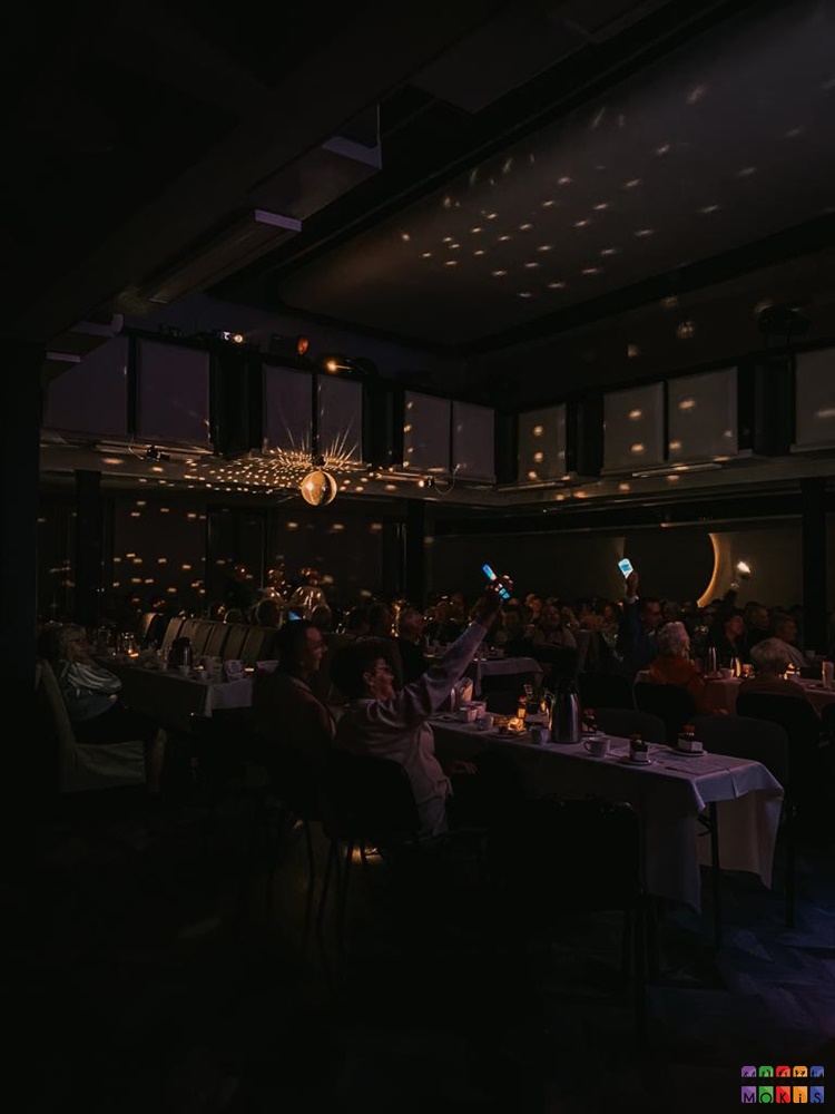 Zdjęcie przedstawiające sale widowiskową z włączoną dyskotekową kulą. W ciemni widać siedzących ludzi przy stolikach trzymających telefony w rękach