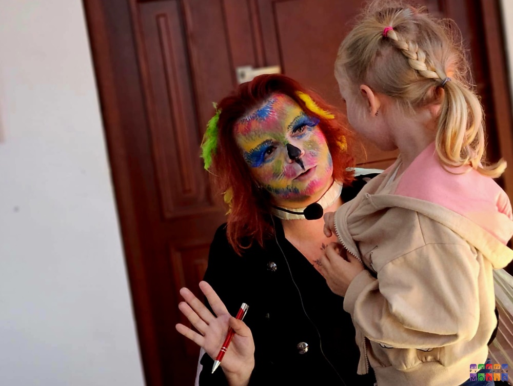 Zdjęcie przedstawia dziewczynkę z warkoczem i osobę z kolorową buzią patrzącą na nią. W tle drzwi