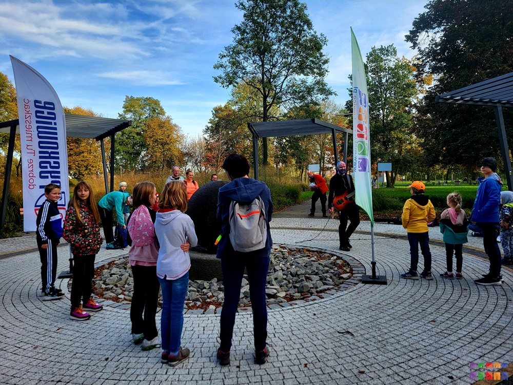 Zdjęcie przedstawia grupę ludzi stojących w kole w parku miejskim. W tle widać drzewa parkowe oraz owiewające flagi z logami MOKiS i Pyskowic