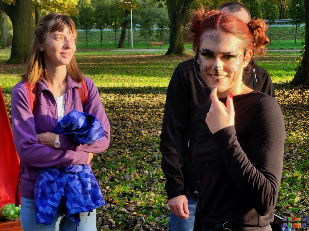Zdjęcie przedstawia troje ludzi stojących w parku. Jedna z nich - kobieta - obrócona i uśmiechnięta w stronę fotografa