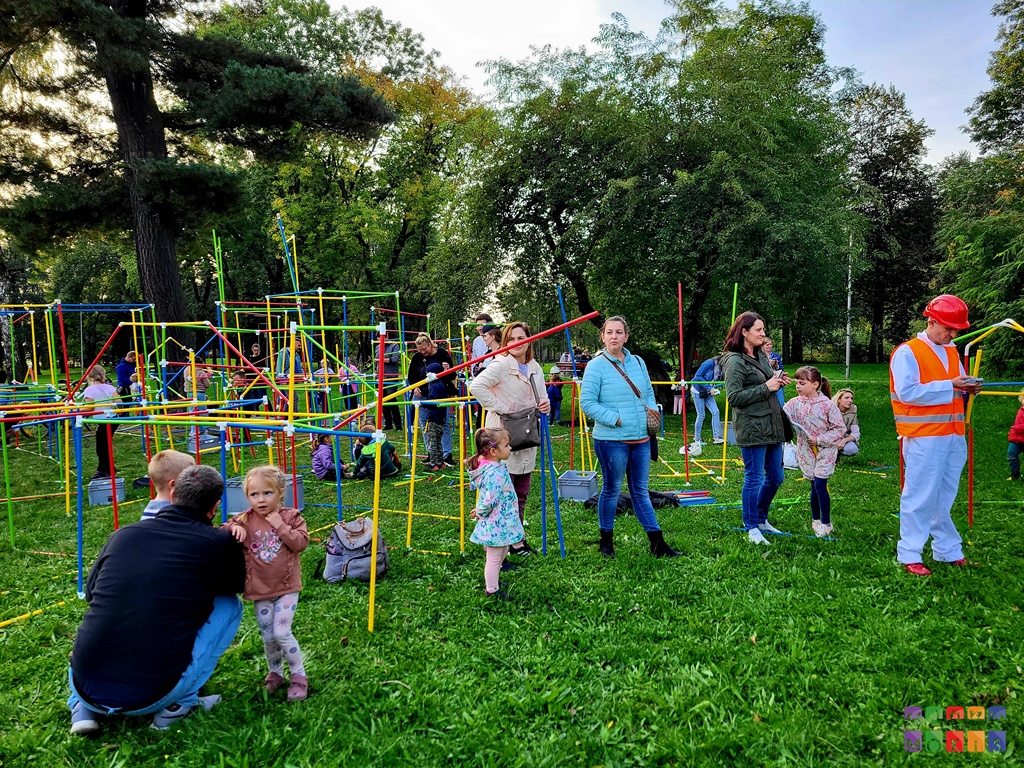 Zdjęcie przedstawiające bawiących się ludzi pomiędzy kolorowymi kijkami w parku. W tle widać drzewa. Z kijków powstaje budowla.