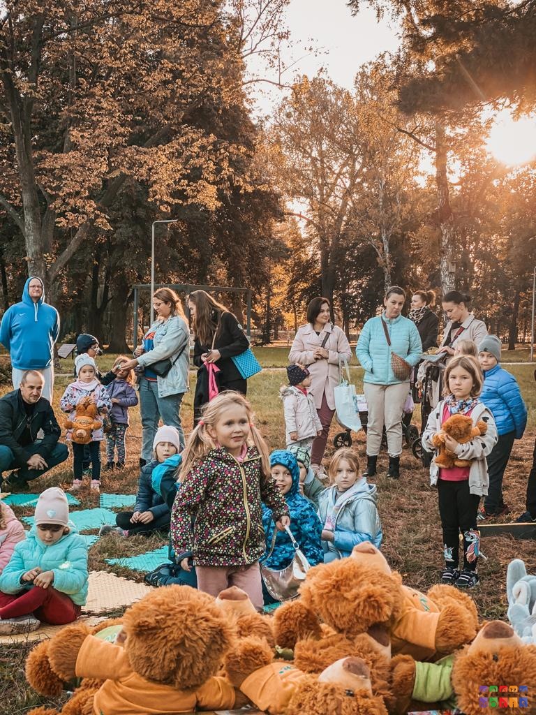 Zdjęcie przestawiające grupę ludzi (dzieci i dorosłych) siedzących i stojących w Parku. W tle widać drzewa parkowe.
