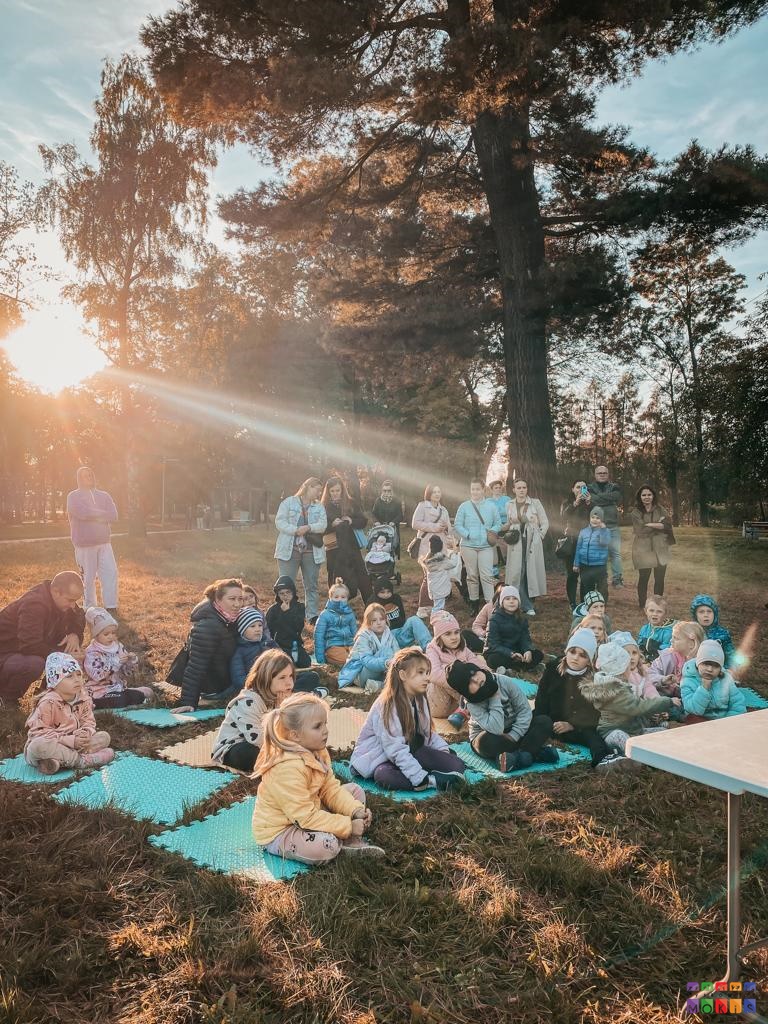 Zdjęcie przedstawiające grupę ludzi a Parku. Jedni z nich dzieci siedzą na piankach natomiast dorośli stoją w tylnej części. W tle widać drzewa parkowe przez, które przebija się słońce.