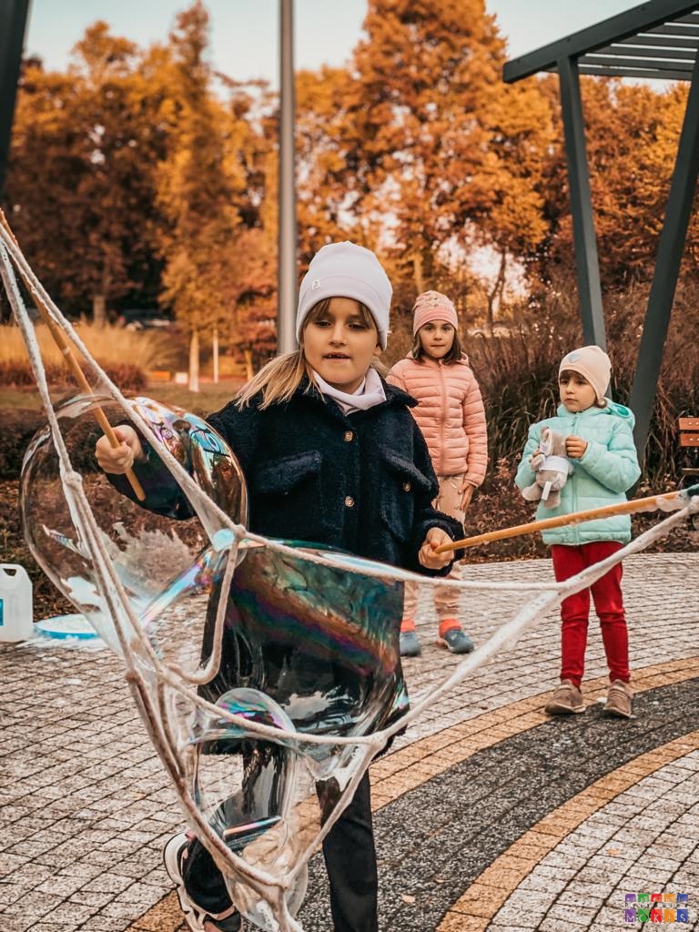 Zdjęcie przedstawiające bawiące się dziewczynki w parku. Na pierwszym planie dziewczyna robi ogromne bańki mydlane sznurkami