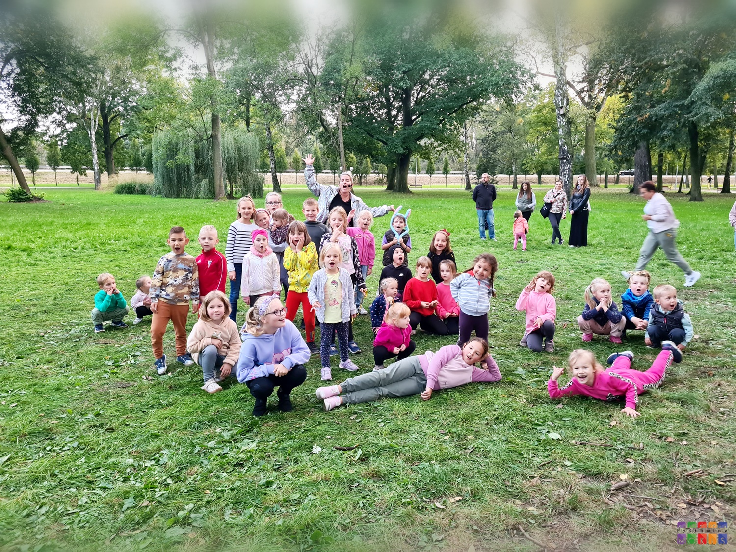 Zdjęcie przedstawiające grupę dzieci siedzących i stojących w parku. W tle widać drzewa parkowe.