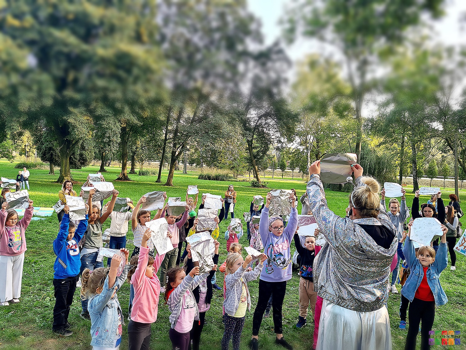 Zdjęcie przedstawiające grupę dzieci siedzących, stojących oraz skaczących w parku. W rękach trzymają srebrne kartki. W tle widać drzewa parkowe.