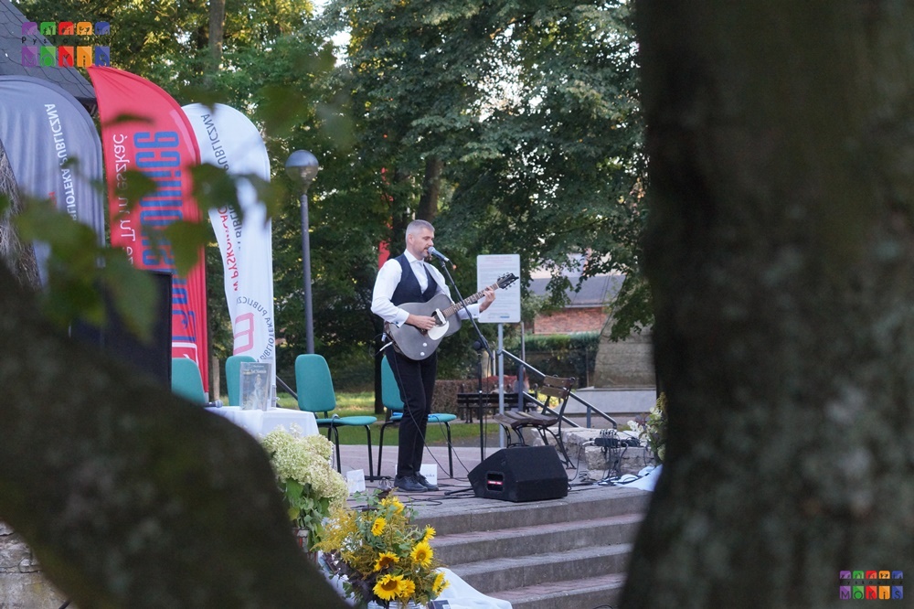 Zdjęcie przedstawiające mężczyznę z gitarą w ręku stojący przy mikrofonie w parku