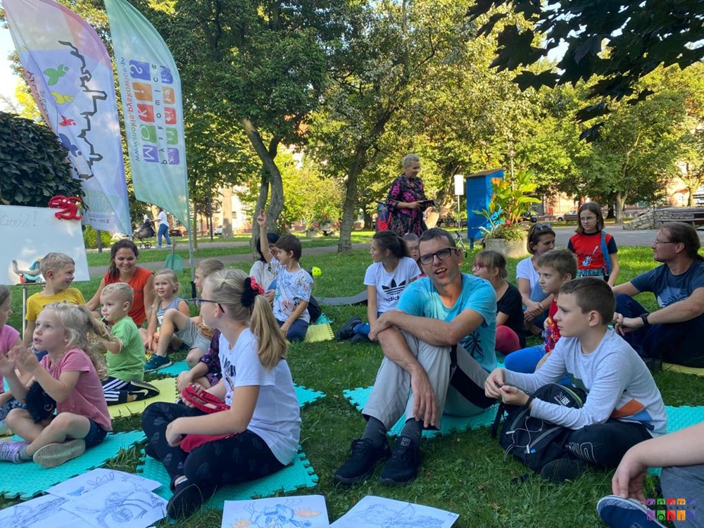 Zdjęcie przedstawiające grup ludzi siedzących na matach w parku. Wśród nich dzieci, młodzież i dorośli. W tle widać drzewa parkowe oraz flagi