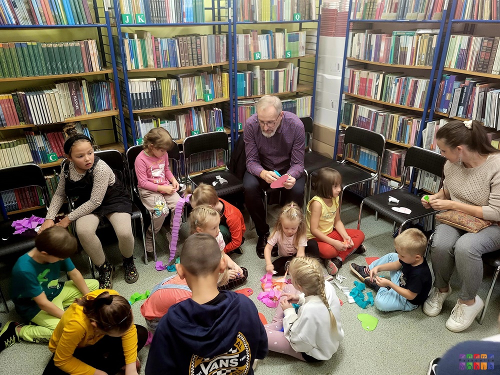 Zdjęcie przestawiające grupę ludzi (dzieci i dorosłych) siedzących na podłodze i krzesłach wśród półek w bibliotece