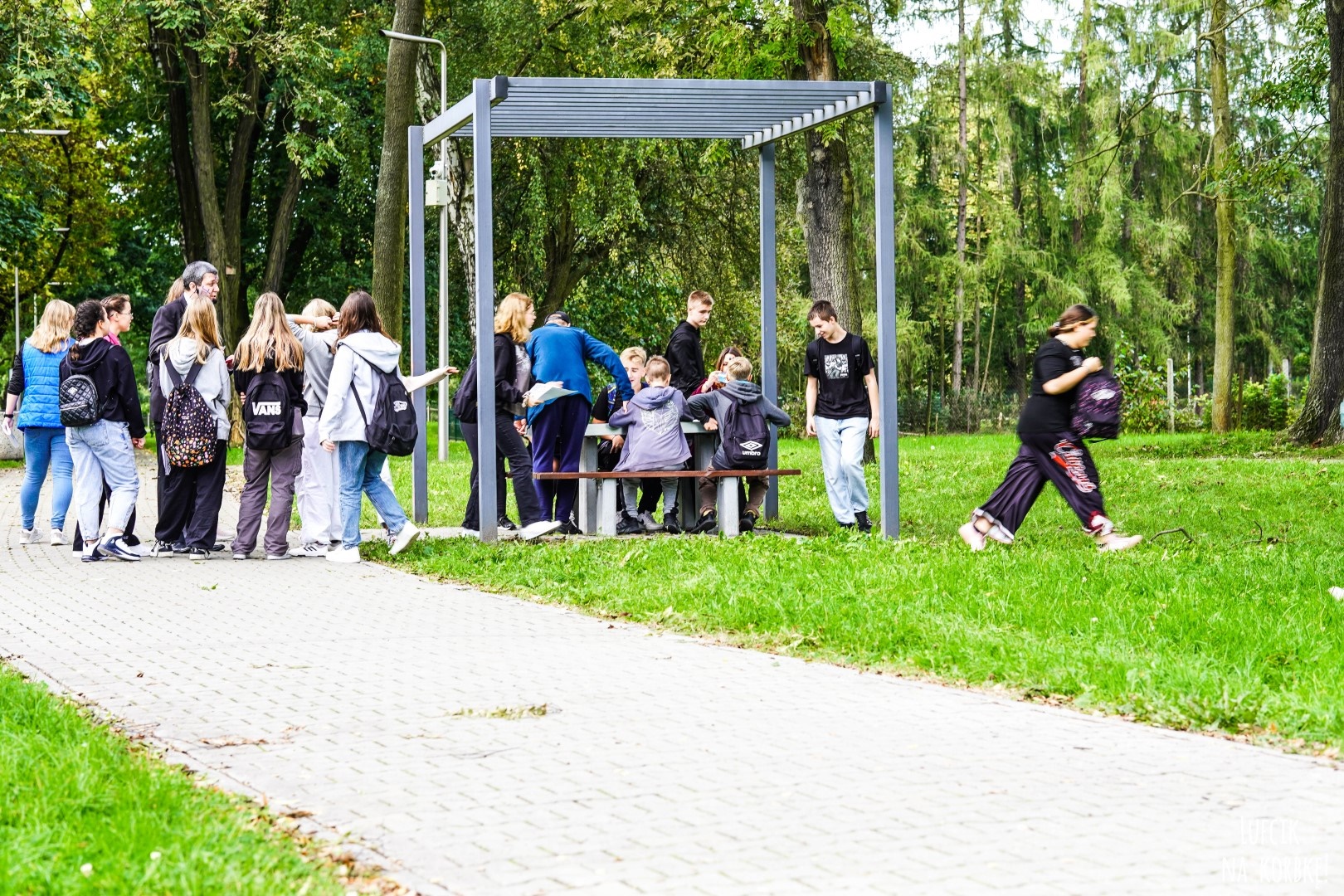 Zdjęcie przedstawiające grupę ludzi w parku. Jedni stoją przy pergoli metalowej drudzy siedzą pod nią przy stoliku. W tle widać drzewa parkowe