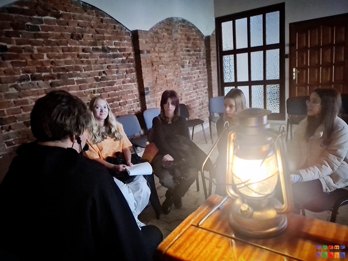Zdjęcie przedstawia siedzące dziewczyny w sali z murami z cegieł. Na pierwszym planie zapalona lampka naftowa. Całość w półmroku.
