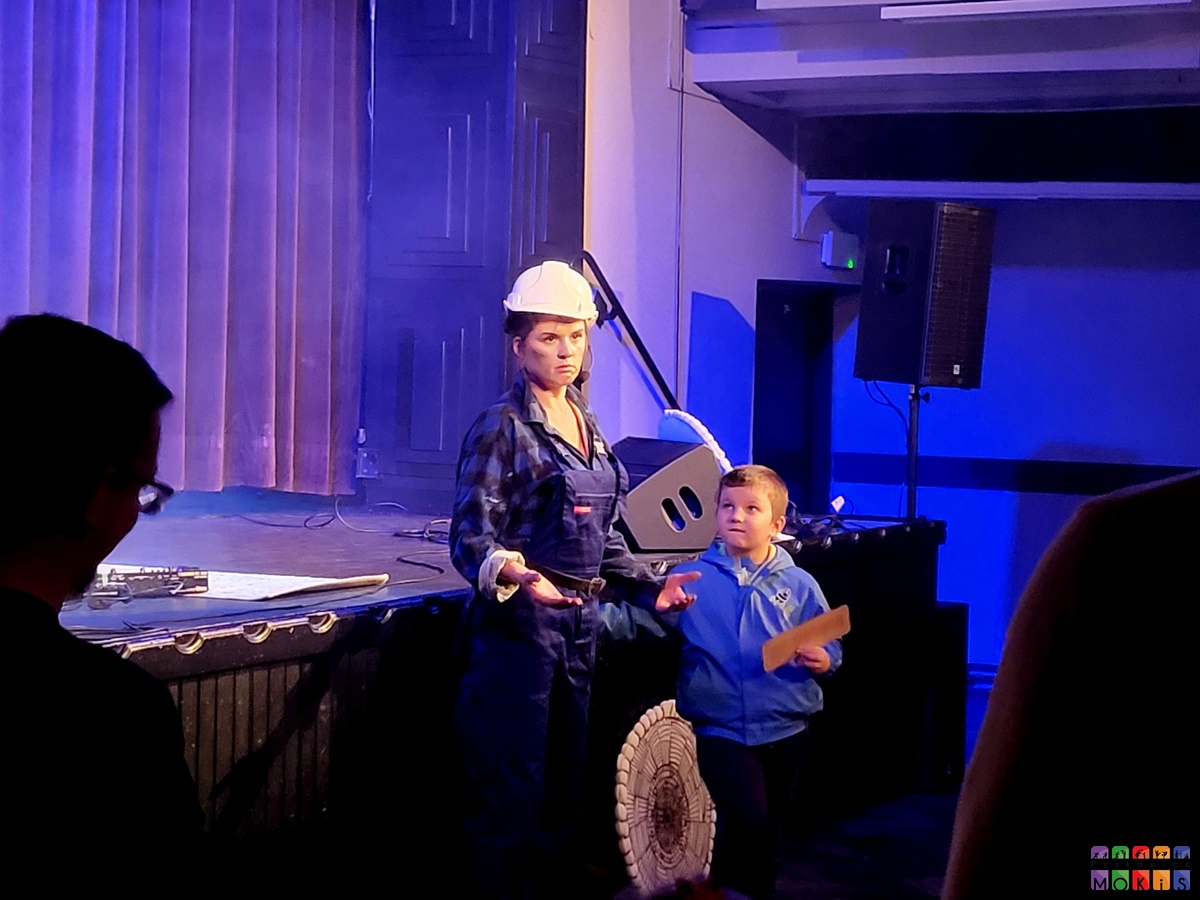 Zdjęcie przedstawia scenę w MOKiS a na przed nią kobieta przebrana w strój roboczy. Odgrywa jakąś rolę. Przy niej stoi chłopiec