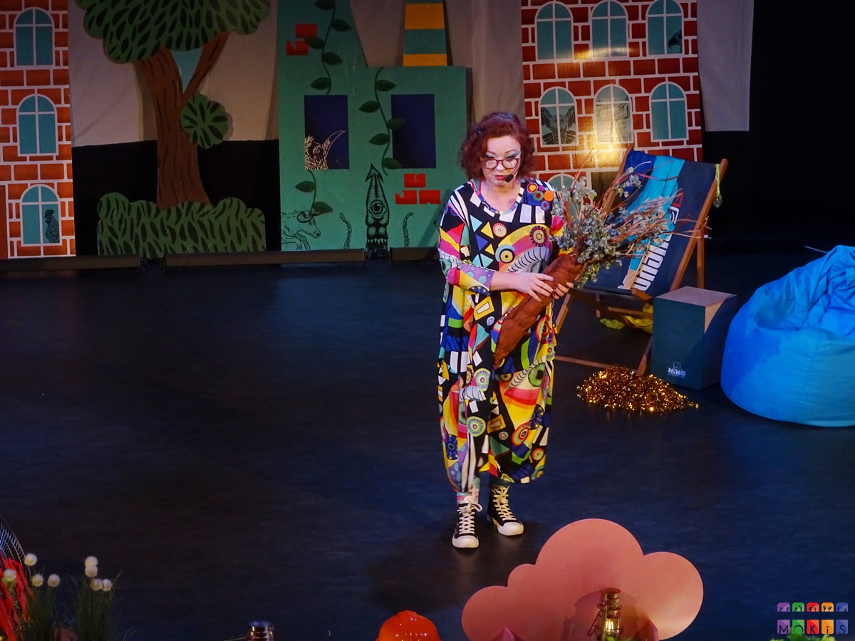 Zdjęcie przedstawiające aktorkę stojącą na scenie, w tle której widać kolorową scenografię. Aktorka ubrana w kolorowy strój.