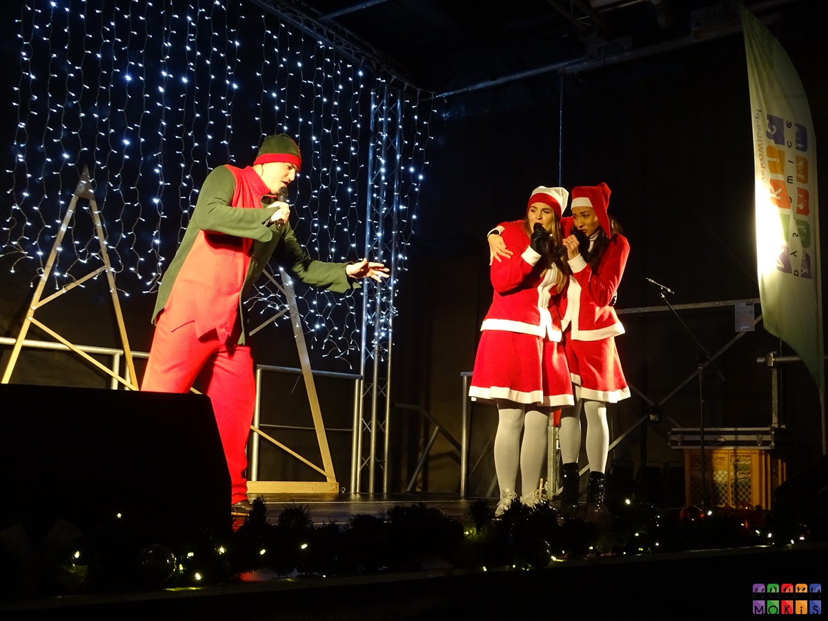 Zdjęcie przedstawiające występy na scenie plenerowej. Dwie kobiety oraz mężczyznę. Wszyscy przebrani świątecznie.
