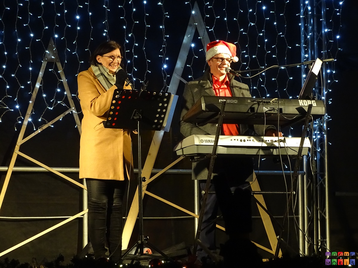 Zdjęcie przedstawiające śpiewającego pana do mikrofonu oraz kobietę na scenie plenerowej. W tle widać choinki drewniane oraz oświetlenie świąteczne.