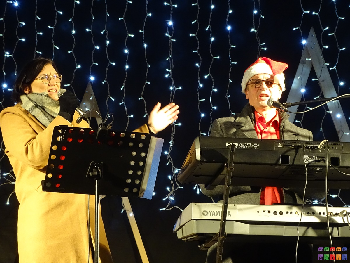 Zdjęcie przedstawiające śpiewającego pana do mikrofonu oraz kobietę na scenie plenerowej. W tle widać choinki drewniane oraz oświetlenie świąteczne.