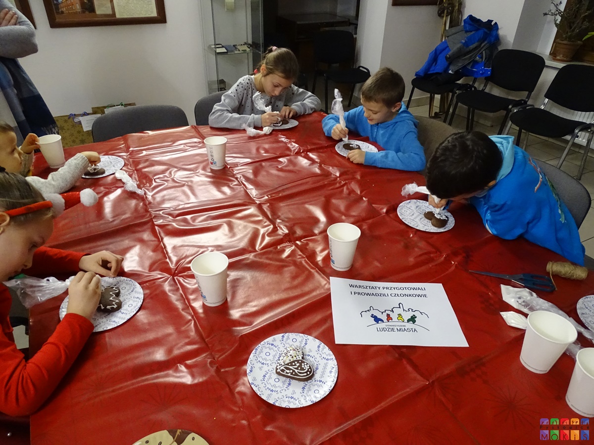 Zdjęcie przedstawia siedzących dzieci przy czerwonym stole i lukrujących pierniki