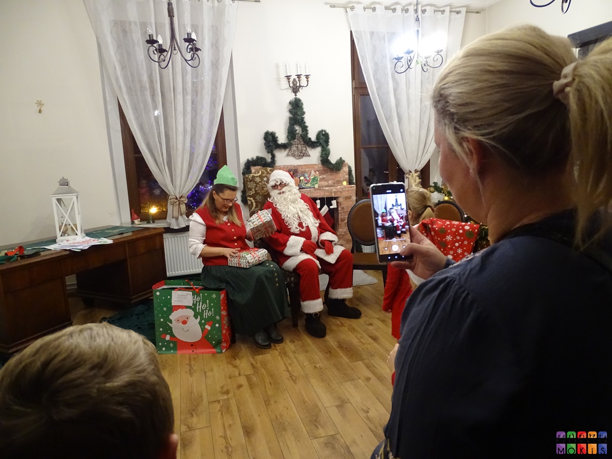 Zdjęcie przedstawia elfa i Mikołaja siedzących na krzesłach a na pierwszym planie widać telefon którym ktoś robi im zdjcie