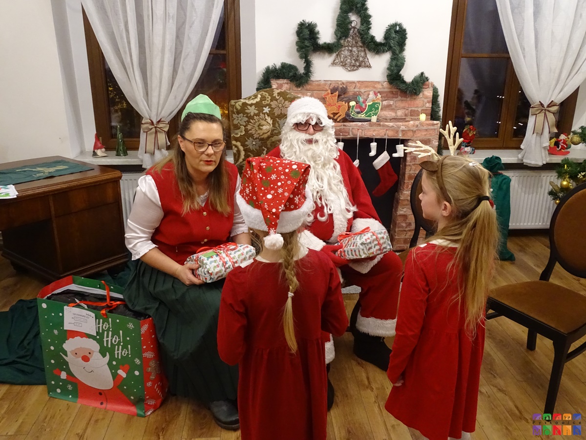 Zdjęcie przedstawia przekazywanie prezentów dzieciom przez Mikołaja i elfa