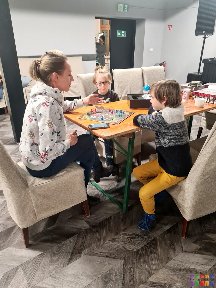 Zdjęcie przedstawia siedzącą kobietę z dziećmi przy stole na którym rozłożona jest gra planszowa.